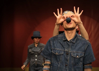 A performer circling the eyes of another performer wearing a black clown nose.