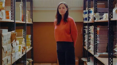 Une femme en pull rouge se tient entre deux étagères recouvertes de manuscrits de livres.