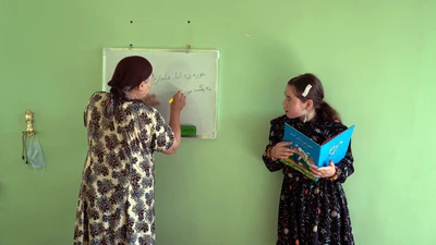 une adulte et une jeune fille, se tiennent devant un tableau blanc fixé à un mur vert. L’adulte écrit en arabe pendant que la jeune fille lit un livre illustré