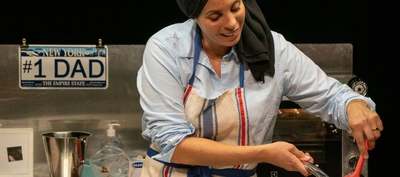 Au non du père La photographie montre une personne derrière un plan de travail de cuisine installé sur une scène ou dans un espace de spectacle. La personne porte une chemise bleu clair, un tablier à rayures verticales bleu, blanc et rouge, ainsi qu’un tissu noir couvrant la tête et tombant sur une épaule.
Au premier plan, la personne utilise une spatule rouge au-dessus d’un récipient ou d’une préparation situé hors du cadre inférieur. Les mains sont occupées à une activité culinaire.
Derrière le plan de travail se trouve une plaque décorative ressemblant à une plaque d’immatriculation américaine. Le texte visible est : “#1 DAD”, avec “NEW YORK” en haut et “THE EMPIRE STATE” en bas.
Autour de la personne se trouvent plusieurs objets de cuisine : un bol métallique, une bouteille de savon liquide transparente, un cadre photo posé sur le plan de travail et un four ou appareil de cuisson en acier inoxydable. Une lumière chaude provenant du four éclaire légèrement l’arrière-plan.
Le décor est sobre, avec un fond noir qui met en valeur la personne et les accessoires. L’ensemble évoque un spectacle mêlant cuisine, récit personnel et représentation théâtrale.