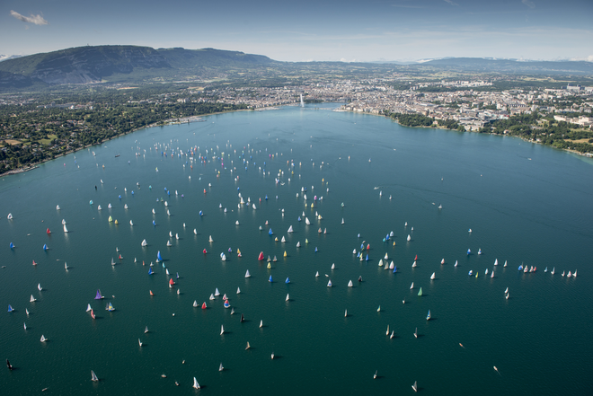 Sur cette image, on aperçoit une partie du lac Léman avec, au fond, la ville de Genève. On distingue également le jet d'eau et les montagnes qui entourent la ville. De nombreux bateaux à voile parsèment le lac, ajoutant une touche dynamique à ce paysage pittoresque.