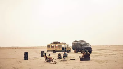 Un desert, deux voitures et plusieurs personnes assises sur des chaises et un canapé devant les voitures. Il y a également deux grosses enceintes pour du son.