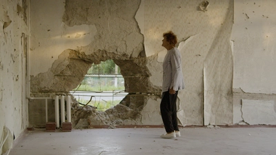 Une femme de profil lève la tête vers le mur abîmé en face d'elle. Le mur à sa droite présent un grand trou.