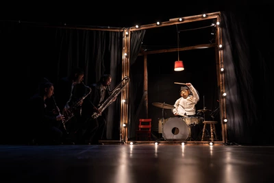 Trois musiciens qui jouent des cuivres, et une marionnette qui joue de la batterie.