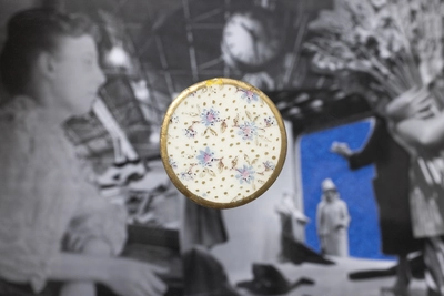 Un bouton en céramique au fond blanc avec des décors floraux et à la bordure dorée est disposé sur une image en noir et blanc et légèrement floue représentant des femmes.