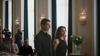 Deux femmes, une en costume, l'autre en robe, se tiennent debout côte à côte. Dans le fond, des personnes assises et deux personnes debout avec des caméras regardent dans la direction des deux femmes.