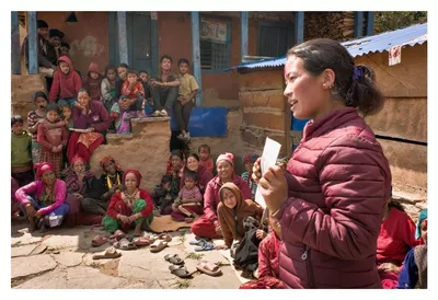 Another World Is Possible – When Communities Make a Difference | Meet Our Local Programme Coordination Teams Une séance de sensibilisation de l’organisation partenaire Sahakarmi au Népal en 2025. On voit, de côté, une femme népalaise qui tient une fiche dans ses mains. Elle est en train de parler à une assemblée communautaire dont on voit quelques membres, femmes, hommes et enfants, assis dans un cercle autour d'elle au sol et sur des escaliers d'un bâtiment au fond de l'image.