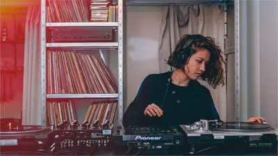 Une femme mix sur des platines de DJ dos à une étagère recouverte de CDs et vinyles.