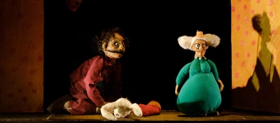 La photographie montre une scène de théâtre de marionnettes éclairée par une lumière chaude. Au centre, sur un plateau en bois, se trouvent deux marionnettes de taille moyenne.
À gauche, une marionnette porte un vêtement rouge foncé à rayures. La marionnette est agenouillée au sol et porte de grandes lunettes rondes. À droite, une seconde marionnette est vêtue d’un costume vert turquoise volumineux et possède de très grandes oreilles blanches qui s’étendent de chaque côté de la tête.
Entre les deux marionnettes repose un objet en tissu blanc évoquant un petit animal ou une peluche, à côté d’un bonnet rouge. Le décor est encadré par deux panneaux verticaux jaunes ornés de petits motifs roses.
À droite de l’image, un éclairage projette sur le panneau jaune l’ombre d’une autre marionnette ou silhouette, visible uniquement sous forme d’ombre. L’arrière-plan est très sombre, ce qui met en valeur les personnages et les objets situés au premier plan.
L’ensemble évoque un spectacle jeunesse mêlant marionnettes, narration et univers fantastique. Aucun texte n’est visible dans l’image.