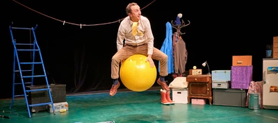 Une scène de théâtre est aménagée avec de nombreux objets du quotidien. Au centre, une personne est assise à califourchon sur un grand ballon jaune de gymnastique. La personne porte une chemise claire, un pantalon beige et des chaussures marron.
À gauche de la scène se trouve une échelle bleue. Un fil à linge est tendu en hauteur d’un côté à l’autre de l’espace, avec plusieurs pinces à linge accrochées. Derrière la personne, un porte-manteau supporte plusieurs vêtements colorés.
À droite, un ensemble d’objets est empilé : valises, boîtes, tiroirs, un petit meuble de chevet, une paire de bottes rouges, une lampe et divers coffres de rangement. Tout au fond à droite, un petit réfrigérateur est décoré de cartes postales ou d’images.
Le sol est vert et l’arrière-plan est constitué d’un rideau noir. L’éclairage de scène met en valeur les objets aux couleurs vives, notamment le ballon jaune, l’échelle bleue et les bottes rouges. Aucun texte n’est visible dans l’image. L’ensemble évoque un univers théâtral ludique et inventif, conçu à partir d’objets familiers.
