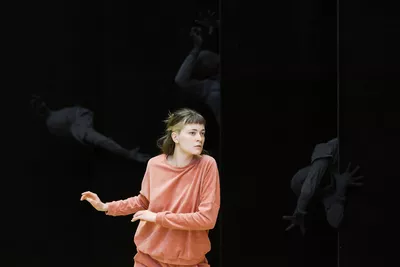 Une jeune femme porte un survêtement couleur saumon. À l’arrière-plan, la scène est sombre, mais on distingue plusieurs personnes vêtues de blanc.