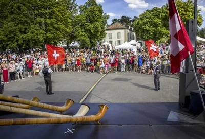 L'image est une photographie d’une célébration de la Fête nationale suisse à Genève en plein air, par une journée ensoleillée. Au premier plan, plusieurs cors des Alpes, longs instruments en bois, sont posés devant une scène. Deux hommes en costume traditionnel brandissent de grands drapeaux suisses rouges avec la croix blanche, face à une large foule rassemblée en demi-cercle. Les spectateurs, debout, comprennent des familles, des enfants et des personnes de tous âges. En arrière-plan, on aperçoit des arbres feuillus, une grande maison ancienne et plusieurs tentes blanches de type événementiel, probablement pour des stands ou animations. L’ambiance est festive et communautaire, typique des célébrations du 1er août à Genève.