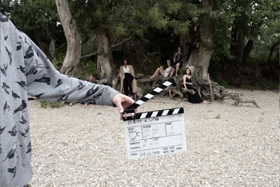 Anti-Nymphs | Gli appunti di Anna Azzori | Projection + conversation Photo d'un tournage dans la nature avec en premier plan un bras tenant un clap de film et en arrière plan des femmes en robes noires assises sur les racines d'un arbre.