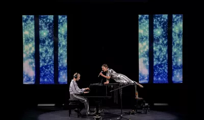 TRILOGIE DE LA GUERRE Un homme joue du piano, un casque sur les oreilles. Devant lui, une femme est allongée, un micro à la main. Tous deux portent des vêtements argentés. Deux panneaux lumineux sont placés de chaque côté de la scène