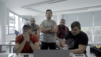 Cinq hommes dans une salles. Quatre d'entre eux sont assis, de part et d'autre de l'homme au milieu qui est debout.