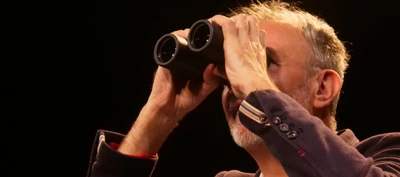 Audience information Gros plan d’une personne sur une scène sombre, éclairée par une lumière chaude. La personne porte une veste foncée avec des boutons visibles aux poignets et tient une paire de jumelles noires devant les yeux à l’aide de ses deux mains.
Le cadrage est serré sur le haut du corps et le visage est en grande partie masqué par les jumelles. On distingue des cheveux courts et une barbe grisonnante. Les bras sont relevés pour maintenir les jumelles en position d’observation.
L’arrière-plan est entièrement noir ou très sombre, sans élément de décor visible, ce qui attire l’attention sur la personne et l’objet qu’elle tient. La lumière met en valeur les contours des mains, des jumelles et du visage.
Aucun texte n’est visible dans l’image. L’ensemble évoque une situation d’observation ou de recherche, dans un contexte théâtral ou de conférence sur scène.