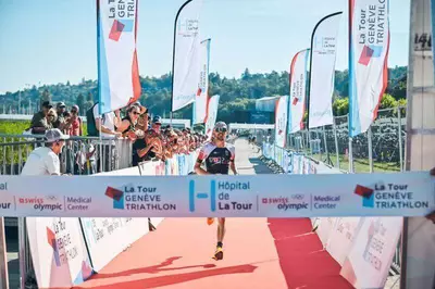 Sur l'image, on voit l'arrivée du Tour Genève Triathlon avec un coureur arrivant seul. De chaque côté, une foule l'encourage et des drapeaux portant le nom de l'événement sont visibles. Un tapis rouge est déposé sur le sol à l'arrivée. Le ciel est bleu, et en arrière-plan, on aperçoit des arbres verts.