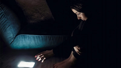 Une femme est assise accroupie. Elle tends sa main vers son téléphone, seule source de lumière dans la pièce.