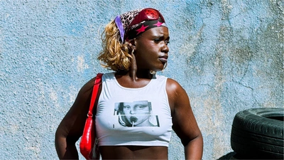 Une femme avec un tshirt représentant une femme avec une sucette pose devant un mur bleuté et des pneus sur la droite de l'image.