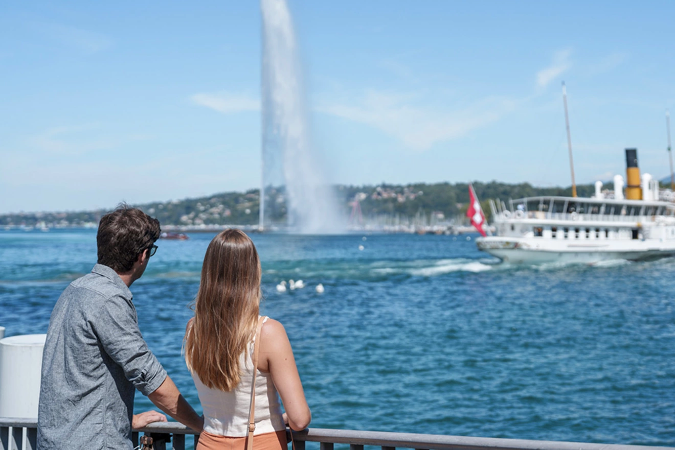 Two people admiring the Jet d’Eau and a passenger boat on Lake Geneva.