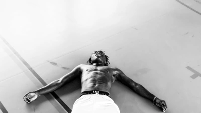 Un homme est allongé sur le dos, les bras ouverts, sur le sol d'un gymnase. La photo est en noir et blanc.