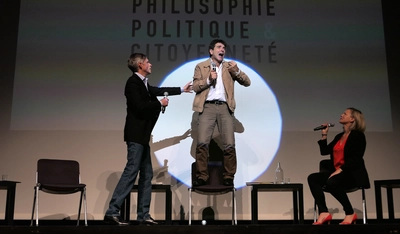 The Republic by Platon Un intervenant très engagé debout sur une chaise face à l'audience parle dans un micro et deux de ses collègues essaient de le calmer.