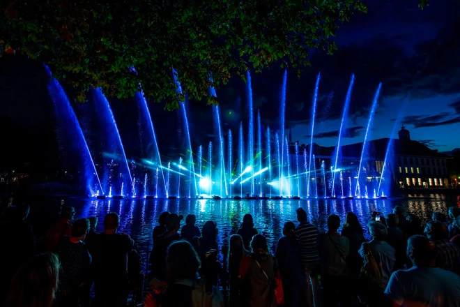 Foule regardant de hauts jets de fontaine illuminés en bleu et vert, avec des faisceaux laser traversant le ciel, sur le lac de Genève de nuit