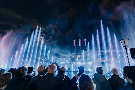 Crowd photographing tall white illuminated fountain jets rising into the night sky, with flags and a lamppost in the foreground