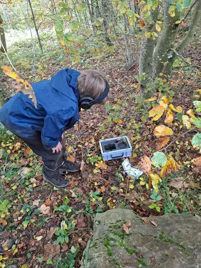 Dans une forêt, un enfant avec un casque sur les oreilles regarde vers le sol. Un équipement est posé au sol.