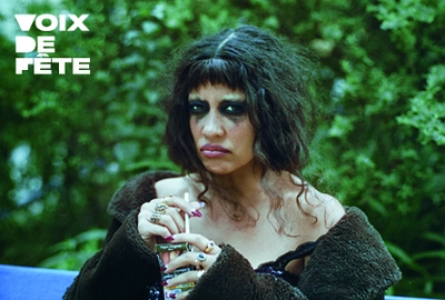 Un portrait de l'artiste Gildaa, les yeux grimés de noir, un verre à la main, assise sur un banc avec un fond de verdure.