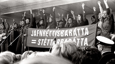 Des femmes sur une estrade en face d'une foule lèvent le poing en brandissant une affiche. L'image est en noir et blanc.