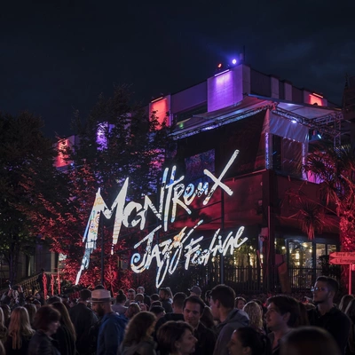 L’image montre une scène de nuit animée au Montreux Jazz Festival. Une foule dense de festivaliers se tient devant un bâtiment moderne illuminé de lumières colorées violettes, rouges et bleues. Le logo du festival, écrit dans une typographie artistique, est projeté en grand au centre de l’image. L’ambiance est festive, avec des jeux de lumière sur les arbres et des palmiers en pot qui complètent le décor.