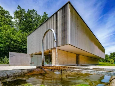 Between Heritage and Renewable Energy: A Guided Visit of Les Berges de Vessy Bâtiment moderne de la Maison du Futur à Vessy, avec fontaine au premier plan et arbres en arrière-plan.