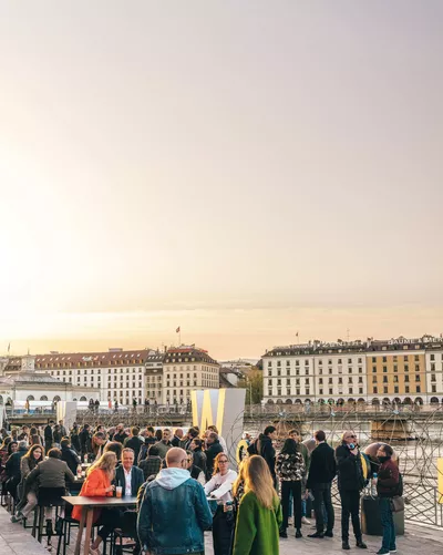 Photographie d’un événement en plein air au bord du lac Léman, à Genève. De nombreuses personnes discutent autour de tables hautes, près d’installations métalliques décoratives, avec les bâtiments du quai des Bergues en arrière-plan, sous un ciel de fin de journée.