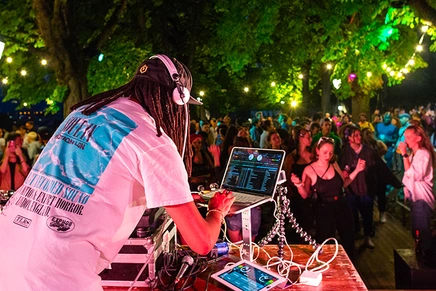 A DJ performing at a laptop and turntables under string-lit trees, with a crowd dancing outdoors at night