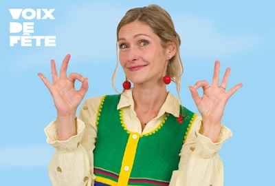 Une image de GiedRé avec un gilet vert sur fond bleu clair. Elle sourit et fait des OK avec les doigts de ses mains.