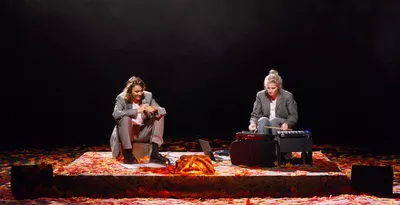 VOIR CLAIR AVEC MONIQUE WITTIG Deux comédiennes sont assises sur scène. L'une avec un micro dans les mains, l'autre bat le rythme avec des baguettes. Le sol est recouvert de feuilles mortes rouges et oranges.