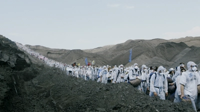 Dans un paysage rocailleux et gris, une longue file de personne en tenue de protection blanche défilent.