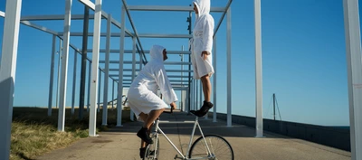 Photographie prise en extérieur sous un ciel bleu dégagé. Au centre de l’image, deux personnes vêtues de peignoirs blancs à capuche et de chaussures noires interagissent avec un vélo blanc.
Une personne est accroupie sur les pédales du vélo, maintenant l’équilibre. L’autre personne se tient debout sur le guidon et semble légèrement surélevée au-dessus du vélo. L’ensemble évoque une figure d’équilibre ou une acrobatie.
La scène se déroule sur une allée en béton bordée d’herbe. Autour des personnes se dresse une structure géométrique composée de nombreux cadres métalliques blancs formant une succession de rectangles qui s’étendent en profondeur. Ces lignes créent un effet de perspective marqué vers l’arrière-plan.
La lumière est vive et naturelle, soulignant le contraste entre les vêtements blancs, le vélo blanc, les structures métalliques et le bleu intense du ciel. Aucun texte clairement lisible n’apparaît dans l’image.
L’ensemble donne l’impression d’une performance artistique ou circassienne mêlant équilibre, mouvement et interaction avec le vélo dans un décor architectural minimaliste.