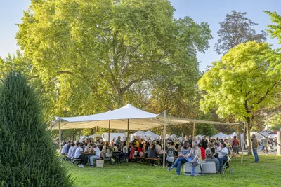 L'image montre une grande tente blanche, installée sur une vaste pelouse verte. Sous cette tente ouverte, de nombreuses personnes sont assises sur des bancs autour de longues tables en bois, partageant un repas.