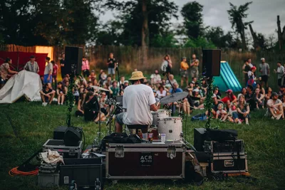 PIZ PALÜ Festival L'image met l'accent sur un batteur de dos. Il fait face à une foule attentive assise dans l'herbe. Le matériel de sonorisation entoure l'artiste, créant une scène improvisée au cœur de la nature.