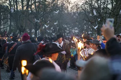 La fête de l'Escalade 2026 Il y a un groupe de personnes vêtues de costumes historiques, principalement des hommes et des enfants, portant des chapeaux à larges bords et des vêtements anciens. Certains portent des torches enflammées, qui ajoutent de la lumière à la scène au crépuscule. En arrière-plan, on peut voir des arbres dénudés, avec des oiseaux blancs qui semblent s'envoler. L'atmosphère est festive et dynamique, avec des gens de tous âges participant à l'événement, certains prenant des photos. L'ensemble évoque une ambiance historique et culturelle, marquée par des couleurs chaudes créées par la lumière des torches.