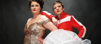 DIVA Syndicate Portrait de deux personnes posant côte à côte devant un fond sombre uni. Les deux personnes regardent vers l’objectif et occupent le centre de l’image.
À gauche, une personne porte une robe longue et brillante recouverte de paillettes dorées. La robe possède des manches tombantes sur les épaules. Une main est posée sur la hanche.
À droite, une autre personne porte une veste de sport rouge vif avec de larges bandes blanches sur les épaules et les manches. Un bras est appuyé sur l’épaule de la personne située à gauche, tandis que l’autre main repose sur la hanche.
Les lèvres des deux personnes sont maquillées en rouge, créant un contraste marqué avec leurs vêtements. Au premier plan, dans la partie inférieure de l’image, un large voile ou tissu blanc translucide occupe une partie de l’espace et masque légèrement le bas des silhouettes.
L’éclairage est dirigé vers les personnes, mettant en valeur les reflets de la robe à paillettes et les couleurs vives de la veste rouge, tandis que l’arrière-plan reste sombre. Aucun texte n’est visible dans l’image.
L’ensemble évoque une photographie promotionnelle de spectacle, avec une esthétique théâtrale, élégante et volontairement contrastée.
