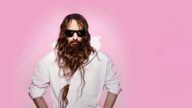 Photo de Sébastien Tellier cheveux longs et lunettes de soleil sur fond rose