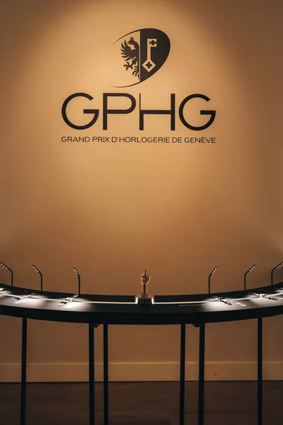 Grand Prix d’Horlogerie de Genève- GPHG 2026 L’image montre un mur beige avec le logo du GPHG – Grand Prix d’Horlogerie de Genève inscrit en grand noir. Au centre, sous le logo, se trouve une vitrine semi-circulaire noire avec un éclairage discret, présentant des montres de prestige. Au milieu de cette vitrine trône un petit trophée doré en forme de main levée. L’ambiance est élégante, sobre et muséale, mettant en valeur l’excellence de l’horlogerie.