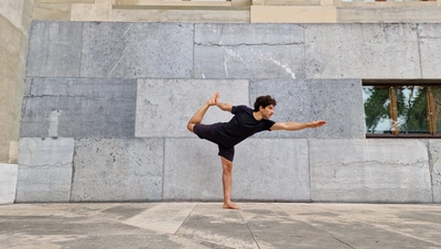 MEG x Groove’N’Move : Yoga Lo-Fi Professeur de yoga faisant une posture de yoga où il tient son pied en l'air devant un mur de brique gris.