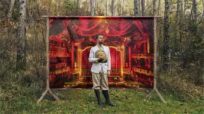 Un homme dans une forêt se tient devant une toile représentant une salle de théâtre dans les teintes rouges / or.