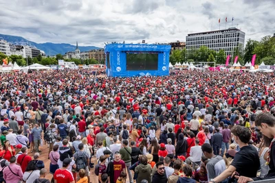 Une foule dense de milliers de personnes, majoritairement vêtues de rouge, est rassemblée en plein air devant un grand écran bleu marqué « Fan Zone Genève 2024 ». L’ambiance est festive. Des tentes blanches bordent les côtés, et des montagnes sont visibles en arrière-plan sous un ciel nuageux. On aperçoit aussi des drapeaux, des bâtiments et une église au loin.