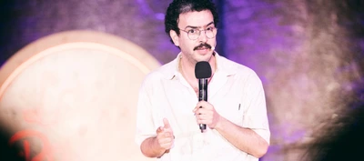 Yacine Nemra is weak Une personne se tient sur une scène et parle dans un microphone tenu à la main. La personne porte des lunettes rondes, une chemise claire à manches courtes et un micro-casque fixé près de la joue. Une moustache est visible.
Le cadrage est serré sur le haut du corps. Une main tient le microphone tandis que l’autre est légèrement levée dans un geste d’accompagnement du discours.
L’arrière-plan est flou et éclairé par des lumières violettes et rosées. À gauche de l’image, on distingue une grande forme circulaire claire, partiellement hors champ. L’éclairage met en valeur la personne au premier plan, tandis que le fond reste diffus et peu détaillé.
Aucun texte n’est visible dans l’image. L’ensemble évoque une représentation sur scène, une conférence ou un spectacle d’humour devant un public.