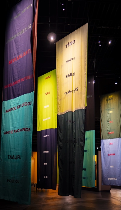 Oeuvre faite de long tissus coloré qui descendent au sol. L'artiste, Bocar Niang, évoque des mots, des griots modernes. Les mots sont en français et en wolof.